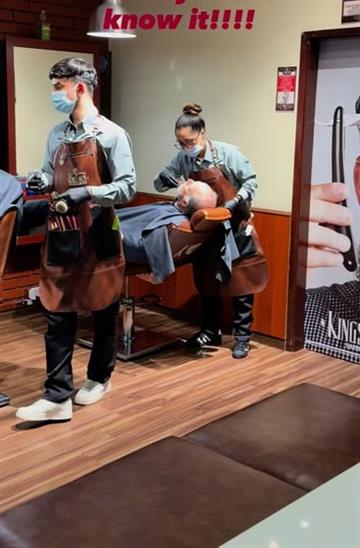 Dos barberos de KingsMen con delantal de cuero atendiendo a un cliente