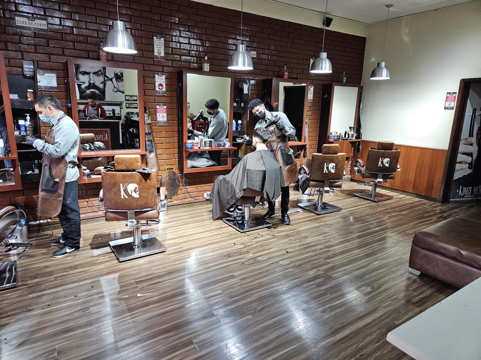 Salón de KingsMen: muros de ladrillo, piso de madera y sillas de cuero con la K, barberos atendiendo